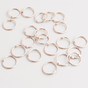 Op de afbeelding: Een verzameling kleine, roségoudkleurige metalen ringen, verspreid over een wit oppervlak. Elke ring is cirkelvormig met een scharnierende sluiting. De ringen worden waarschijnlijk gebruikt voor het maken van sieraden of knutselprojecten.