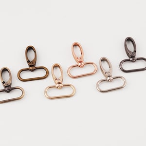 Könnte beinhalten: Sechs Metall-Drehhaken in verschiedenen Ausführungen: Bronze, Roségold, Silber und Dunkelgrau. Jeder Haken hat eine ovale Basis und einen Drehverschluss zum Befestigen. Die Haken sind auf einer weißen Oberfläche angeordnet.
