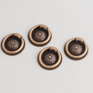 Könnte beinhalten: Vier antike bronzefarbene Schrankgriffe mit einem Blumendesign in der Mitte. Jeder Griff hat einen Ringgriff und eine dekorative kreisförmige Basis. Die Griffe sind auf einer weißen Oberfläche angeordnet.