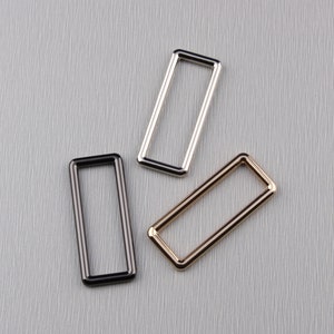 May include: Three rectangular metal rings in black, silver, and gold. The rings are empty in the center and have a smooth, shiny finish. They are arranged on a textured, light gray surface.