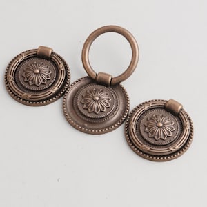 May include: Three antique bronze cabinet pulls. Each pull features a circular design with a central floral motif and a ring handle. The pulls are made of metal and have a vintage aesthetic.
