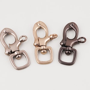 Puede incluir: Tres cierres giratorios metálicos en plata, dorado y bronce oscuro. Cada cierre tiene un gancho con resorte y una base giratoria rectangular. Los cierres están diseñados para sujetarse a bolsos, llaveros u otros accesorios.