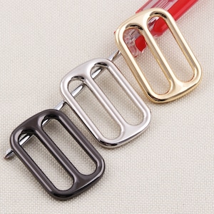 May include: Three metal rectangle slider buckles in black, silver, and gold. Each buckle has a rectangular shape with two parallel bars. The buckles are arranged diagonally on a light beige fabric.