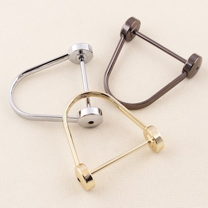 May include: Three metal U-shaped hardware pieces in silver, gold, and dark bronze. Each piece has a circular base and a curved bar, creating a distinctive design. The hardware is arranged on a neutral, textured surface.