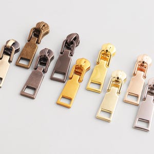 Puede incluir: Una colección de tiradores de cremallera metálicos en varios colores, incluyendo dorado, bronce y plateado. Los tiradores tienen forma rectangular con un agujero en la parte inferior y un lazo en la parte superior.