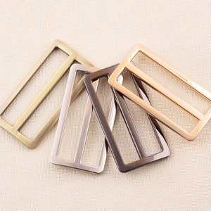 May include: A collection of rectangular metal sliders in various finishes: brass, silver, gunmetal, and rose gold. Each slider has a rectangular frame with three interior bars, suitable for straps or belts. The hardware is displayed on a neutral fabric background.