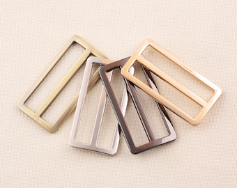 May include: A collection of rectangular metal sliders in various finishes: brass, silver, gunmetal, and rose gold. Each slider has a rectangular frame with three interior bars, suitable for straps or belts. The hardware is displayed on a neutral fabric background.