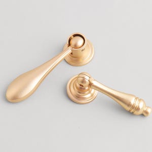 May include: Two gold-colored cabinet pulls with a teardrop-shaped handle and a circular base. The pulls are made of metal and have a smooth, matte finish. The hardware is designed for furniture and home decor.