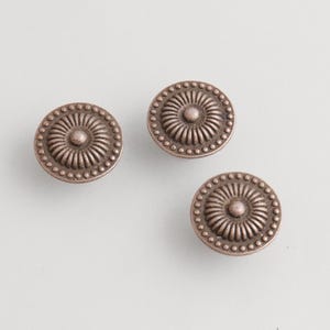 May include: Three antique bronze cabinet knobs with a detailed, circular design. Each knob features a central, raised circle surrounded by radiating lines and a beaded outer edge. The knobs are displayed on a light background.