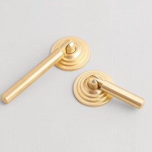 May include: Two gold-colored cabinet door handles. Each handle has a round base with concentric circles and a straight, cylindrical lever. The handles are made of metal and have a matte finish.