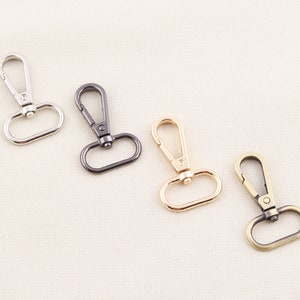 May include: Four metal swivel clasps in silver, gunmetal, gold, and antique brass. Each clasp has a lobster claw hook and a D-ring base. The clasps are displayed on a neutral, textured background.