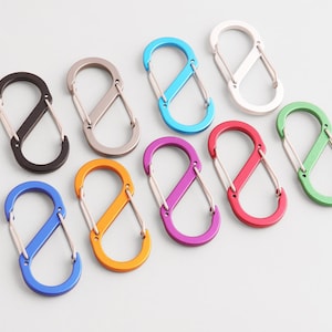 May include: A collection of colorful, double-ended carabiner clips in various colors, including black, silver, blue, orange, purple, red, and green. These metal clips are designed with a unique figure-eight shape and are ideal for attaching items.