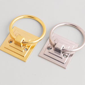 Op de afbeelding: Twee meubelringen, één goudkleurig en één zilverkleurig. Elke ring heeft een vierkante achterplaat met een trapsgewijs ontwerp en een cirkelvormige ringgreep. De hardware wordt getoond tegen een lichtgrijze achtergrond.