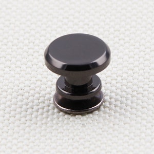 May include: A black cabinet knob with a rounded top and a cylindrical base. The knob is made of a dark, possibly metallic material, and is designed for furniture or cabinetry. The knob is shown against a neutral background.