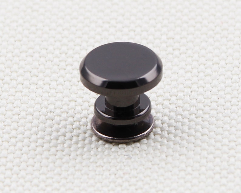 May include: A black cabinet knob with a rounded top and a cylindrical base. The knob is made of a dark, possibly metallic material, and is designed for furniture or cabinetry. The knob is shown against a neutral background.
