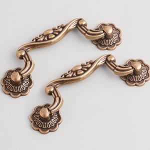 Peut inclure: Deux poignées de tiroir antiques de couleur bronze avec un motif floral. Chaque poignée a une poignée incurvée et des pièces d'extrémité florales décoratives. La quincaillerie est en métal.