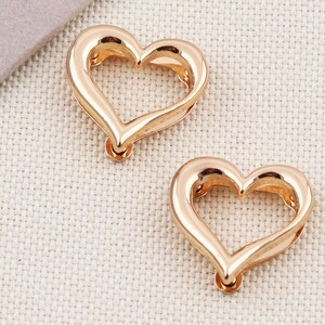 May include: Two rose gold heart-shaped charms. Each charm has a smooth, polished finish and a small loop at the bottom for attaching to jewelry. The charms are displayed on a neutral, textured surface.