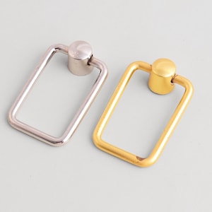 Puede incluir: Dos tiradores de metal para armarios. Uno es de color plateado y el otro dorado. Cada tirador tiene un asa rectangular y un pomo circular. Los tiradores son probablemente para cajones o armarios.