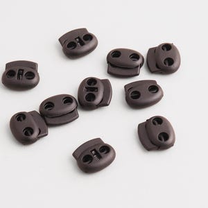 Puede incluir: Un conjunto de topes de cordón marrón oscuro, de forma ovalada con dos agujeros y un mecanismo central. Estos cierres de plástico están diseñados para ajustar cordones y cuerdas, comúnmente utilizados en ropa y accesorios.