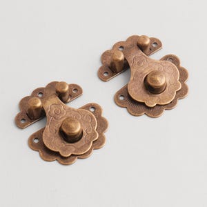 Puede incluir: Dos pestillos antiguos de color bronce con diseños florales decorativos. Cada pestillo tiene un borde festoneado y un botón central para abrir y cerrar. El herraje está diseñado para un armario o una caja pequeña.