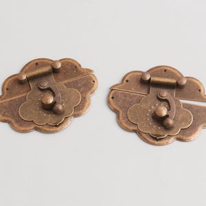 Puede incluir: Dos cierres antiguos de color bronce con forma de nube. Cada cierre tiene un mecanismo de bisagra y un pomo decorativo. El metal tiene un aspecto envejecido, lo que sugiere edad y uso. Estos cierres son probablemente para una caja o cofre.