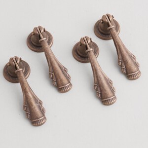 May include: Four antique bronze cabinet pulls with a decorative design. Each pull features a round backplate and a teardrop-shaped handle with intricate detailing, perfect for adding a vintage touch to furniture.