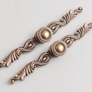 May include: Two antique bronze-colored cabinet pulls. Each pull features a central circular knob with a beaded edge and a decorative, curved handle with ornate leaf and scroll details. The hardware is designed for furniture.