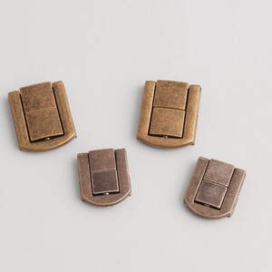 May include: Four antique bronze-colored magnetic clasps. Each clasp has a rectangular shape with a hinged top and a magnetic closure. The clasps are arranged on a light gray surface.