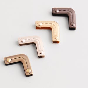 Op de afbeelding: Een assortiment decoratieve metalen hoekbeugels in verschillende afwerkingen, waaronder brons, goud en zilver. Elke L-vormige beugel heeft een decoratieve gestippelde rand en is bevestigd met kleine, ronde klinknagels. De beugels zijn gerangschikt op een lichtgrijze ondergrond.