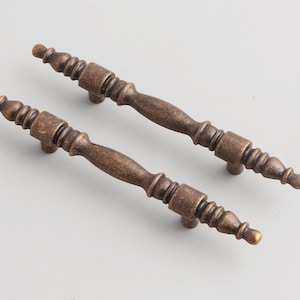 May include: Two antique-style bronze cabinet pulls. Each pull has a central, cylindrical grip with decorative detailing and two mounting posts. The pulls are approximately 13 cm long.