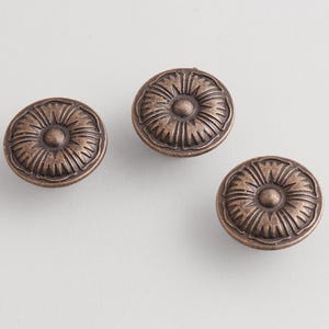 Puede incluir: Tres tiradores de armario de color bronce con un diseño floral. Cada tirador tiene un elemento circular central rodeado de crestas en forma de pétalo. Los tiradores están dispuestos sobre una superficie gris claro.