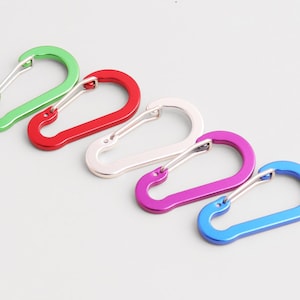 May include: Five colorful carabiner clips in green, red, silver, purple, and blue. These aluminum alloy clips have a spring-loaded gate and are designed for securing items. Ideal for outdoor activities, these clips are lightweight and durable.
