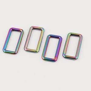 May include: Four rectangular metal rings with a rainbow iridescent finish. The rings are evenly spaced and have rounded corners. The color shifts from blue to green to yellow to pink. The hardware is likely for bags or other accessories.