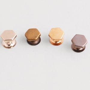 May include: Four metallic cabinet knobs in varying shades of rose gold, bronze, and copper. Each knob has a hexagonal top and a rounded base. The knobs are arranged in a row against a white background.