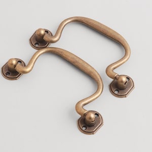 May include: Two antique-style bronze drawer pulls. Each pull has a curved handle and decorative base with screw holes. The hardware is made of metal and has a vintage finish, suitable for cabinets or furniture.