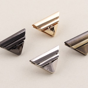 May include: Four triangle-shaped metal clasps in different finishes: gold, silver, black, and antique brass. Each clasp has two raised lines.