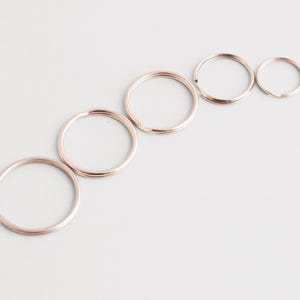 May include: Five rose gold key rings of different sizes are arranged diagonally on a white surface. The key rings are made of metal and have a smooth, shiny finish. The sizes range from approximately 2 cm to 4 cm in diameter.