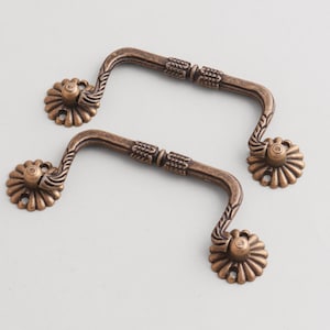 Puede incluir: Dos tiradores de cajón antiguos de color bronce. Cada tirador tiene un asa curva con detalles decorativos y placas de montaje en forma de flor. Ideal para la restauración de muebles o proyectos decorativos.