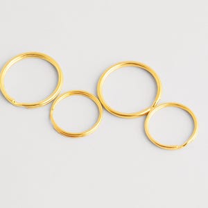 May include: Four gold-colored key rings of varying sizes are arranged on a light gray surface. The rings are circular, with a smooth, shiny finish. The key rings are likely made of metal and are designed to hold keys or other small items.