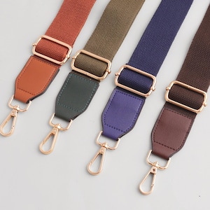 Może przedstawiać: Cztery kolorowe paski do toreb w odcieniach rdzy, oliwkowej zieleni, granatu i brązu. Każdy pasek posiada złotą metalową klamrę i zapięcie, z pasującym skórzanym akcentem w uzupełniającym kolorze.