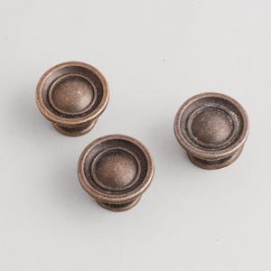 May include: Three bronze-colored cabinet knobs with a circular design. Each knob has a rounded center and a detailed outer ring. The knobs are made of metal and are suitable for furniture hardware.