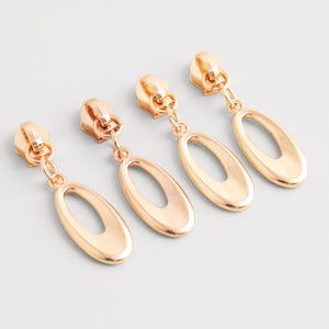 May include: Four gold-colored zipper pull charms with an oval cutout design. The charms are arranged in a row, each with a zipper-shaped top and a dangling oval pendant. The background is a plain, light gray surface.