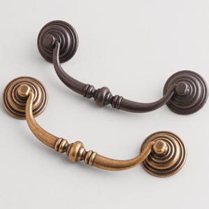 May include: Two decorative drawer pulls. One is bronze-colored, the other is a dark brown. Each pull has a circular backplate with concentric rings and a curved handle. The handles have a decorative, rounded center.