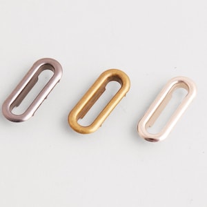Puede incluir: Tres ojales ovalados de metal en diferentes colores: un rosa apagado, un dorado y un oro rosa claro. Estas piezas de ferretería se utilizan para diversos proyectos de artesanía y costura, proporcionando un elemento decorativo y funcional.
