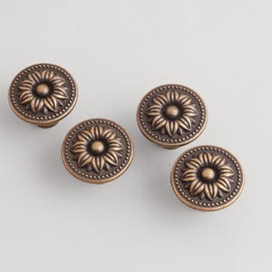 Puede incluir: Cuatro tiradores de armario de bronce antiguo con diseño de girasol. Cada tirador es redondo con un patrón floral detallado en el centro y un borde punteado. Los tiradores son de metal y tienen una estética vintage.