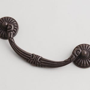 Peut inclure: Une poignée de tiroir en métal de couleur bronze foncé, composée d'une poignée incurvée avec un détail nervuré central et deux plaques de montage circulaires. Chaque plaque présente un motif radial en rayons de soleil avec un petit bouton central.