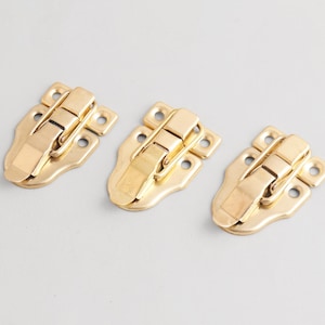以下が含まれることがあります： 3つの金色の金属製の留め金。各留め金は、丸みを帯びたエッジとヒンジ付きのロック機構を備えた長方形のベースを備えています。留め金は、箱や容器を固定するように設計されています。背景は明るいグレーです。