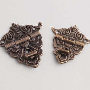 Puede incluir: Dos bisagras ornamentadas de color bronce. Cada bisagra presenta un diseño floral decorativo con detalles enrollados y un pasador central. Las bisagras son probablemente para una caja pequeña o un objeto decorativo.