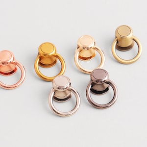 May include: A collection of six metal ring pulls in various finishes: rose gold, gold, bronze, and silver. Each pull features a circular ring attached to a round base. The hardware is designed for use in furniture or craft projects.