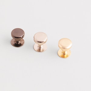 May include: Three metal cabinet knobs in different metallic finishes: dark bronze, rose gold, and gold. Each knob has a rounded top and a circular base, suitable for furniture or decorative projects.